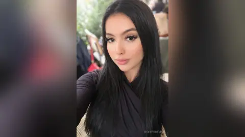 Young woman with long dark hair and glam makeup wearing a black long-sleeve top in a bright indoor setting.