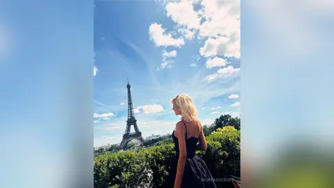 NickyAshcroft poses in a black dress with her blonde hair down, looking out over a view of the Eiffel Tower under a blue sky.