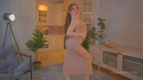 Young woman in a beige midi dress posing in a cozy, warm-lit modern living room.