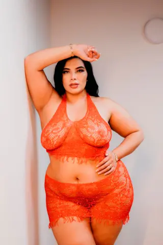 Curvy woman in orange lace lingerie set posing confidently against a neutral wall.