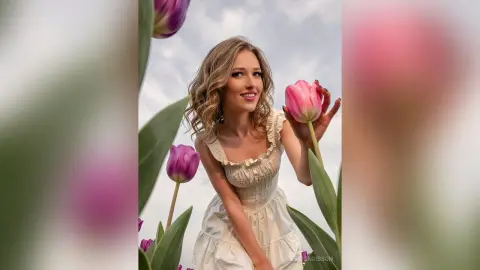 Smiling woman in a white ruffled sundress holding a pink tulip in a dreamy spring garden setting.