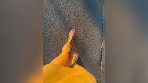 Woman in a mustard yellow dress and strappy sandals walking on a sunlit urban sidewalk.