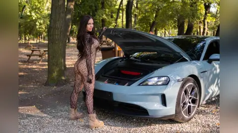 Woman in a patterned bodysuit posing next to a light blue luxury car in a sunny forest setting.