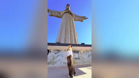 Woman in white top and leopard print skirt standing before the Christ the Redeemer statue under a blue sky.