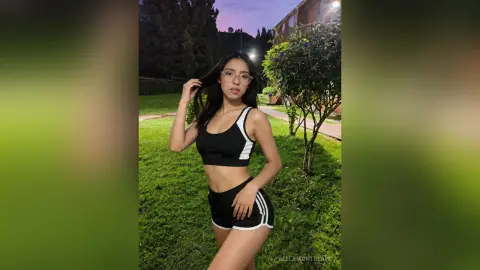 Young woman in black athletic crop top and shorts standing on a green lawn at dusk.
