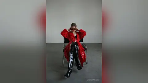 Fashionable woman in a red satin robe and black patent leather boots sitting in a minimalist studio setting.