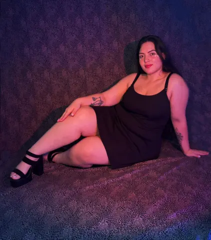 Curvy woman in a black mini dress sitting on a textured purple backdrop with moody lighting.
