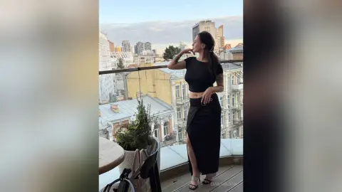 EmilyOlson poses on a balcony overlooking a city, wearing a black short-sleeved crop top and a long black skirt with a high slit.