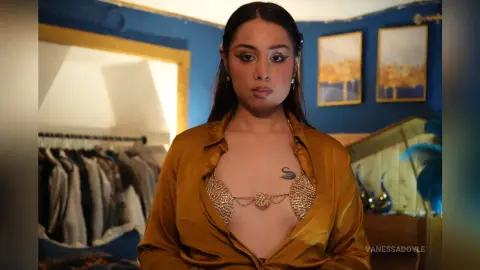 Confident woman in a gold silk shirt and ornate gold bra top posing in a blue bohemian bedroom.