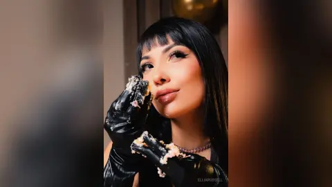 Woman with black bangs wearing black gloves eating messy food in a moody, dimly lit setting.