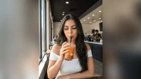 Young woman in a white off-the-shoulder top sipping iced bubble tea in a bright modern cafe.