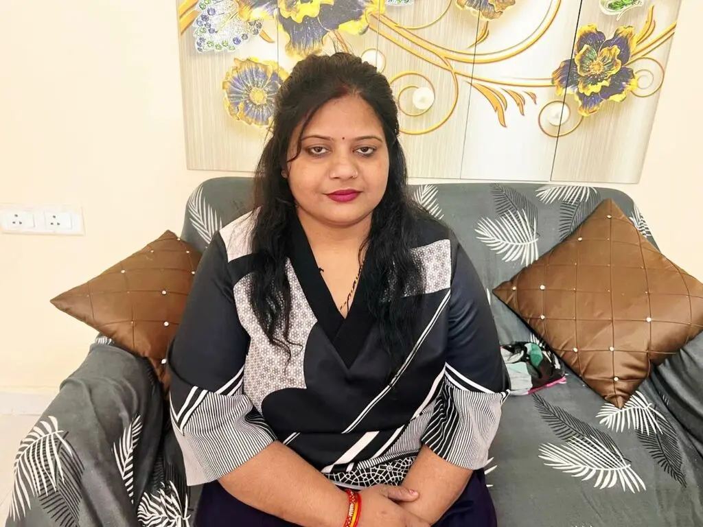Woman in a black and white patterned blouse sitting on a grey patterned sofa in a bright living room.