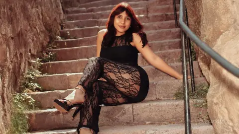 Stylish woman in black lace jumpsuit sitting on stone stairs in a warm, moody outdoor setting.