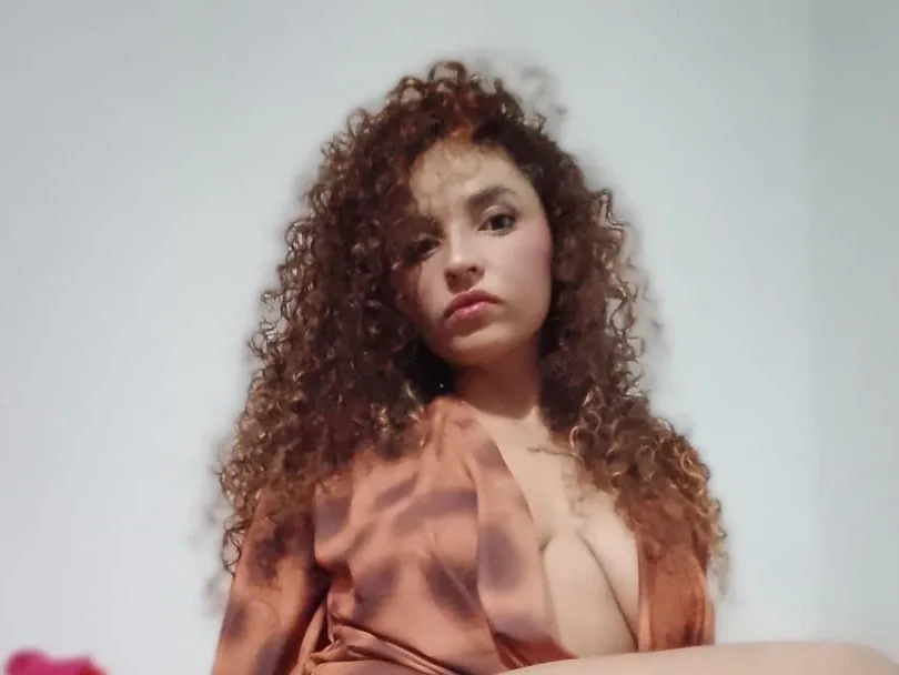 Penelope_Cox wears glasses and a black blazer while looking into the camera with her long curly hair down.