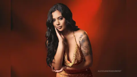 AlessandraDumont poses in a gold sequin dress with her long dark hair flowing over one shoulder against a red background.