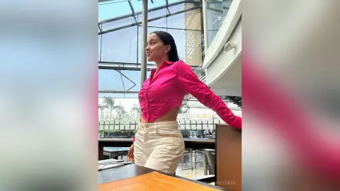 Smiling woman in a pink button-down shirt and white trousers standing in a bright, modern glass building.