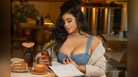 Young woman in a blue denim top reading a book and holding a phone at a cozy cafe setting.