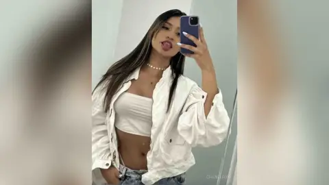 Young woman in a white crop top and open button-down shirt taking a playful mirror selfie in a bright room.