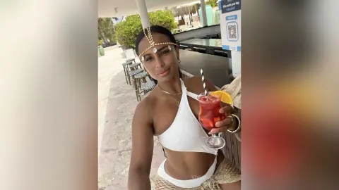 Smiling woman in white bikini and gold headpiece holding a red cocktail at a sunny outdoor tropical resort.