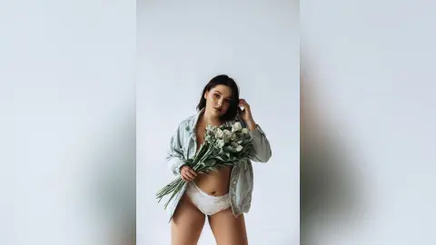 Young woman in an open denim shirt and white lace lingerie holding a bouquet of flowers against a white background.