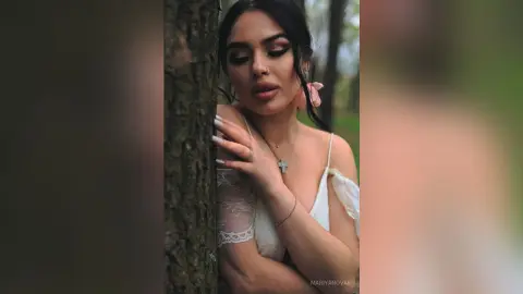 Dreamy portrait of a woman in a white lace dress leaning against a tree in a lush green forest.
