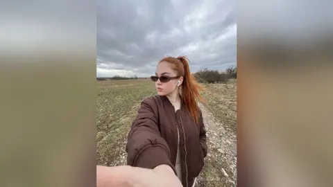 Redheaded woman in a brown bomber jacket standing in a vast field under a cloudy sky.