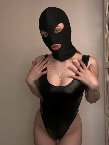 Woman in a black bodysuit and matching face mask posing in a minimalist indoor setting.
