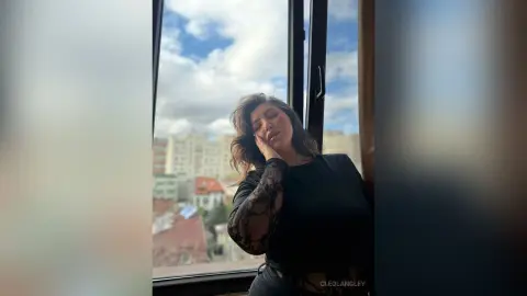 CleoLangley poses with her hand against her face, wearing a black long-sleeved lace top while standing in front of a large window with a city view.