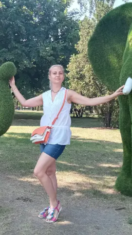 Smiling woman in a white sleeveless blouse and denim shorts posing by green topiary bushes in a sunny park.