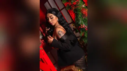 Elegant woman in a black outfit posing near festive Christmas greenery and red decor in a moody setting.