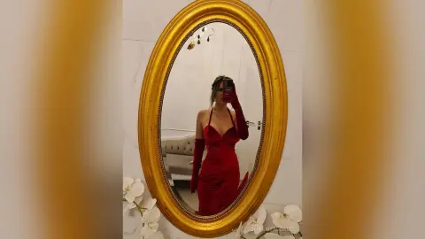 TiffanyAddams poses in a red dress and long red gloves, reflected in a large gold oval mirror.