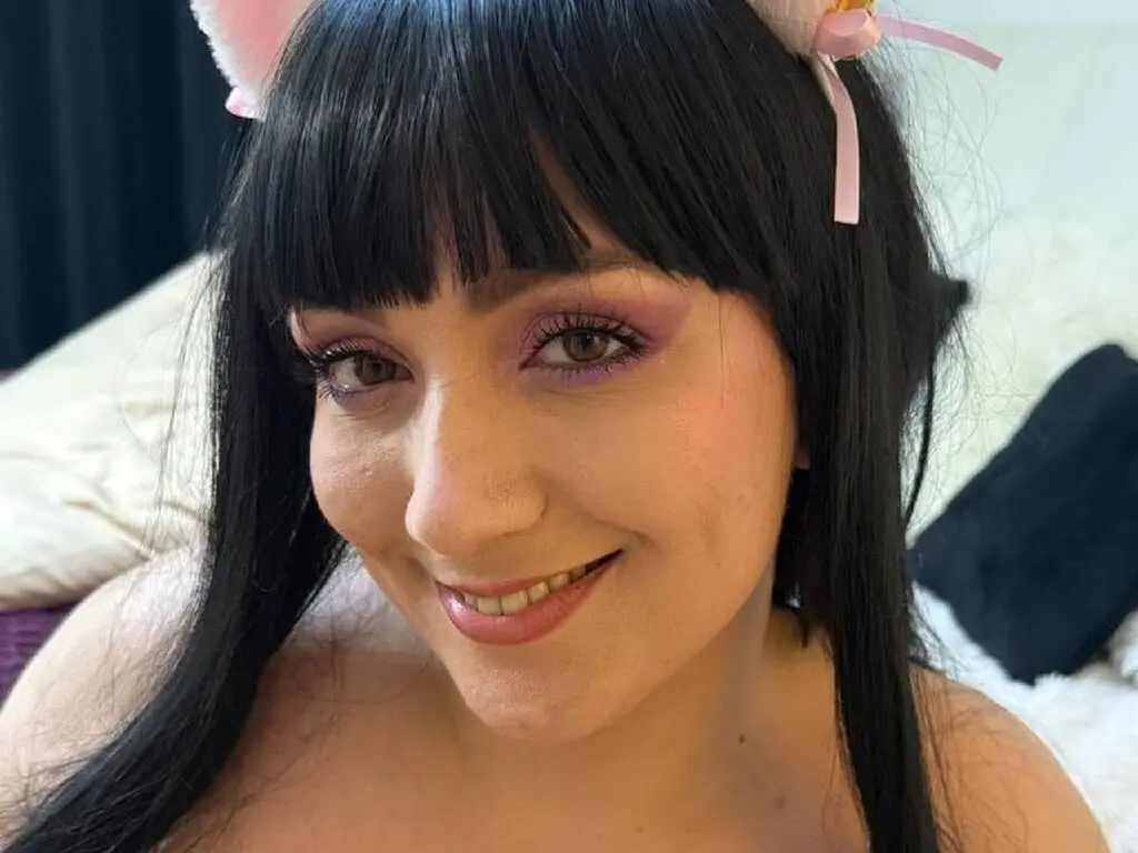 Smiling woman with black bangs and cat ear headband wearing soft makeup in a cozy indoor setting.