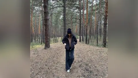 Woman in black winter jacket and jeans walking through a serene pine forest on a cloudy day.