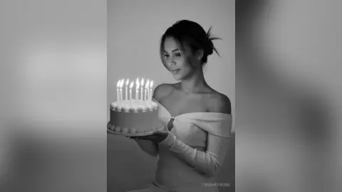 Elegant woman in an off-the-shoulder top holding a birthday cake with lit candles in a moody black and white portrait.