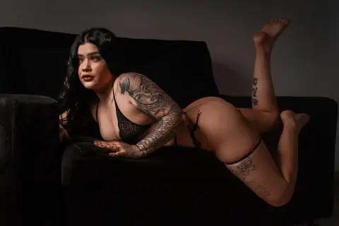 Madisson_Lee20 lies on a black couch wearing black lace lingerie, showing off her tattoos and dark hair.