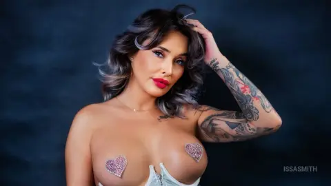 Glamorous woman with wavy hair and arm tattoos wearing heart-shaped glitter pasties in a moody studio setting.