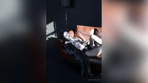 KailaTaylor lies back on a brown leather couch wearing a black lace bra and black stockings, showing off her legs and dark hair.