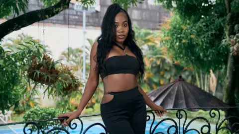 Young Black woman in a black crop top and leggings posing by a garden pool on a sunny day.
