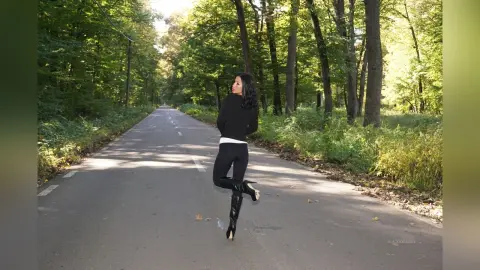 Stylish woman in black outfit and boots walking on a sunny forest road surrounded by lush green trees.