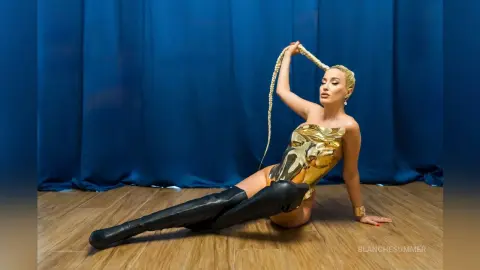 Glamorous woman in a metallic gold bodysuit posing against a blue curtain backdrop.