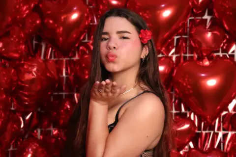 Young woman in black lace top blowing a kiss against a romantic red heart balloon backdrop.