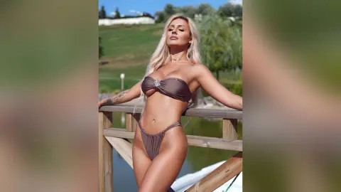 Anita poses outdoors in a brown string bikini, leaning against a wooden railing with her long blonde hair down.