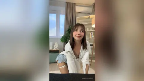Smiling young woman in a white button-down shirt and denim shorts sitting in a bright, modern living room.