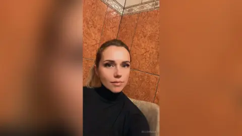 Blonde woman in a black turtleneck sweater posing against a warm terracotta tiled wall in a moody setting.