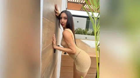 IrinaRussel poses against a wall in a tight, tan mesh mini dress, showing off her long dark hair and slim figure.