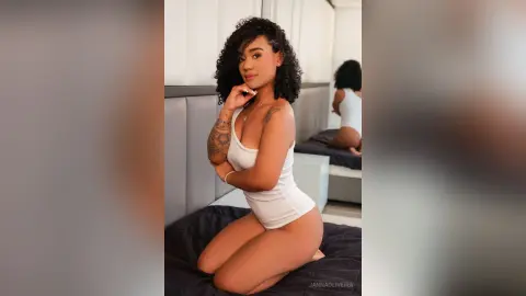 Smiling woman in a white bodysuit sitting on a dark beanbag in a bright, modern bedroom.