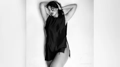 LaurenKaterine poses with her hands behind her head, wearing a short black sleeveless top that shows her legs against a white background.