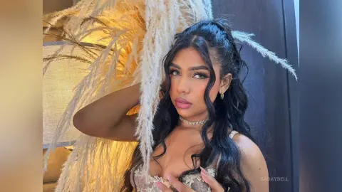 SadayBell poses with long wavy black hair and a lace bra, framed by soft white pampas grass decor.