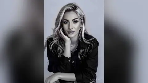 JessycaPage poses with her hand under her chin, wearing a black button-down shirt and showing her long blonde hair.