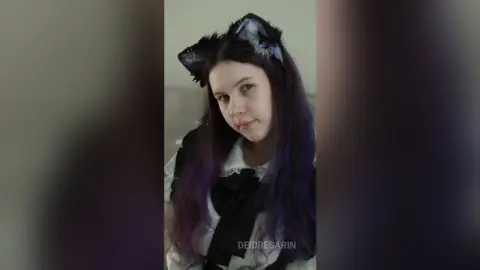 Young woman with purple hair wearing cat ears and a black lace outfit in a soft, moody indoor setting.